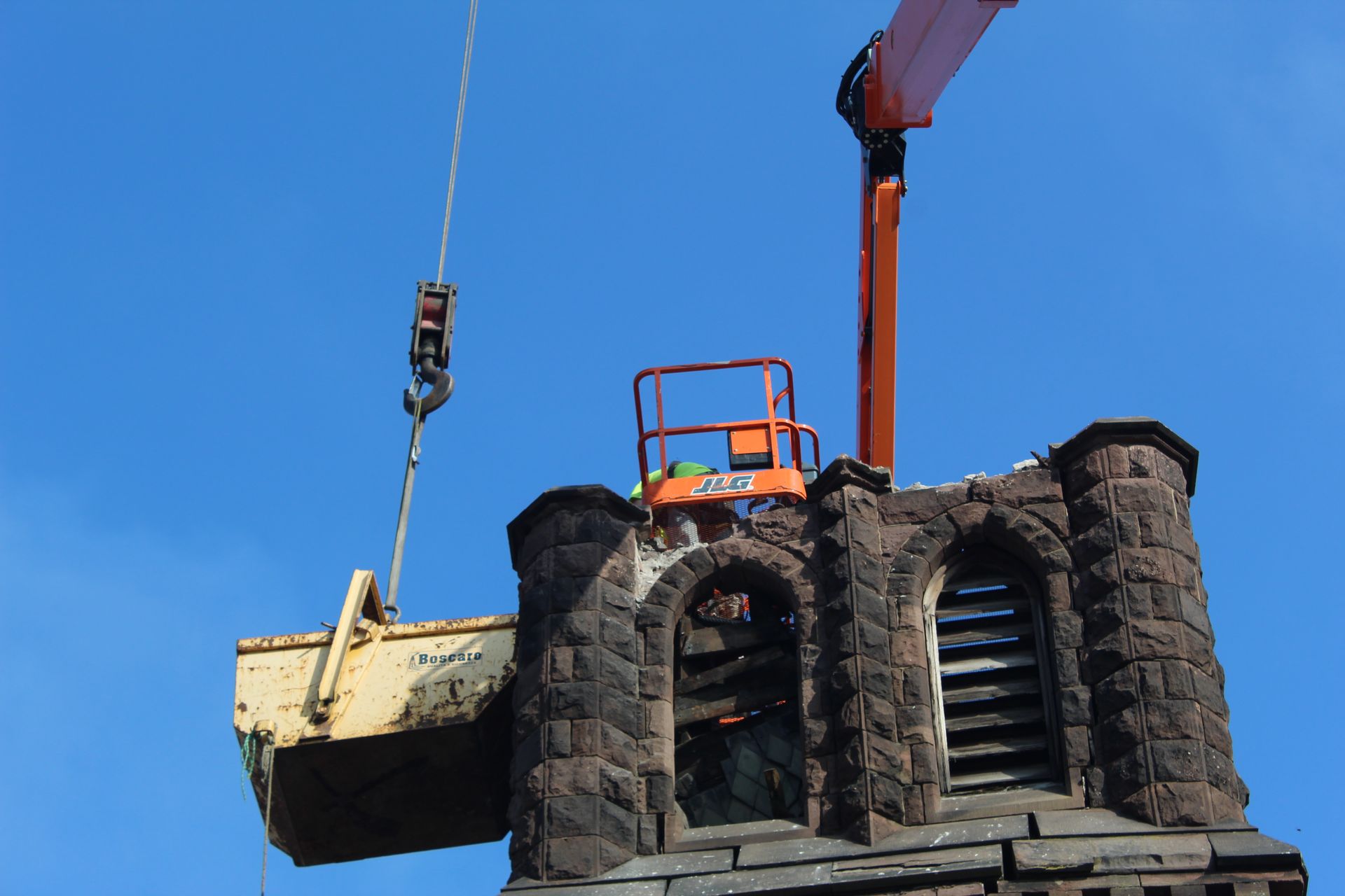 Crane lifting church steeple debris from a damaged stone tower against a bright blue sky.