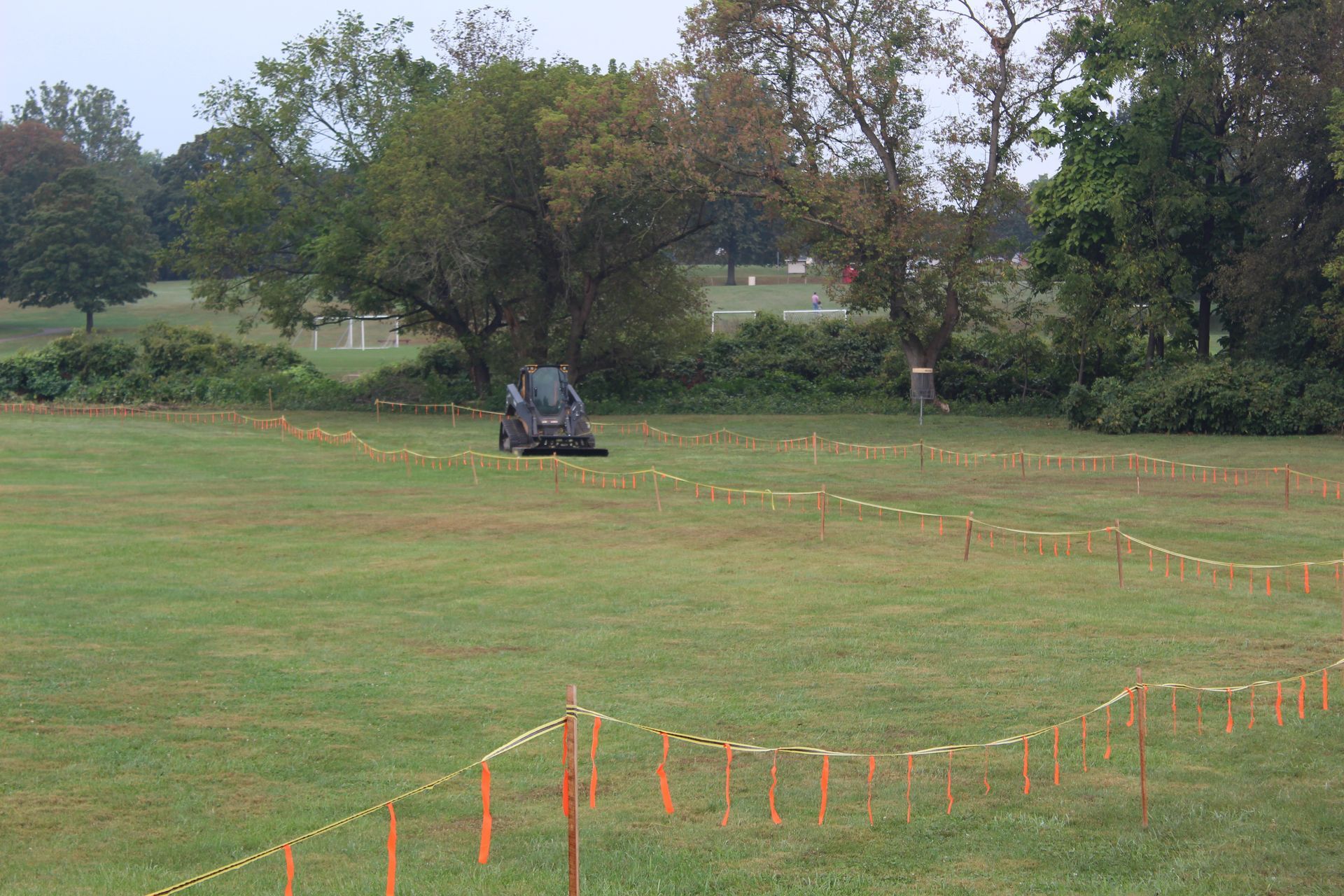A small tractor mowing a grassy field. Orange tape marks a path. Trees and a cloudy sky are in the background.