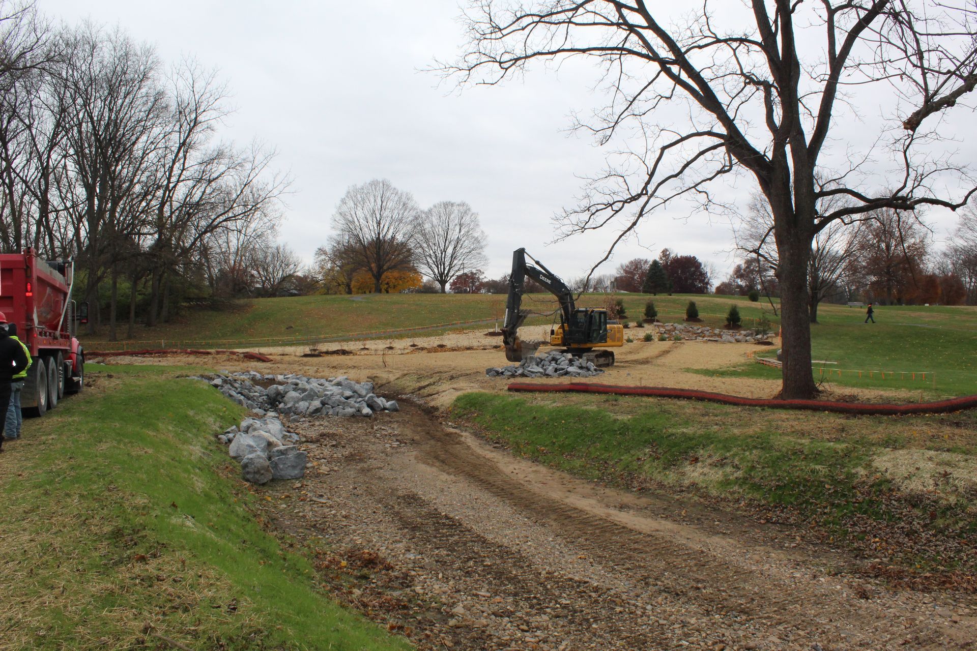 Construction site: Excavator and dump truck near a grassy area and trees on a cloudy day.