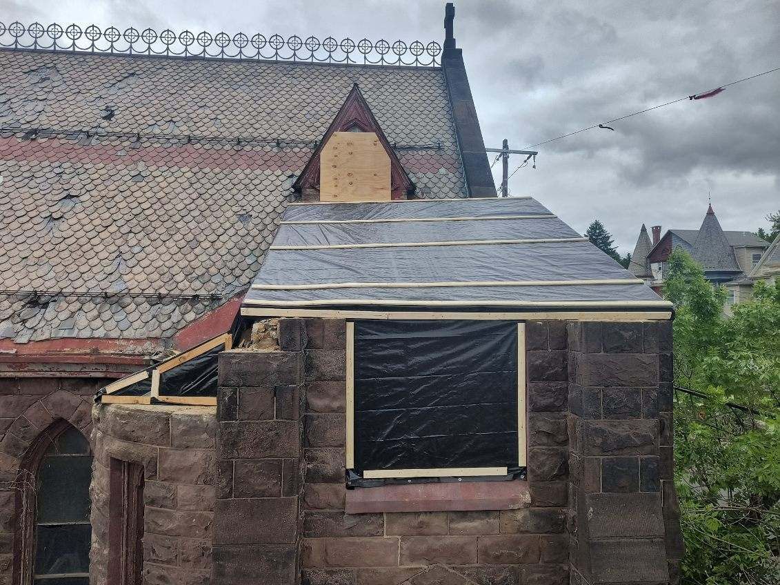 Exterior view of a church under renovation; stone walls, new roof section, plastic-covered window.  