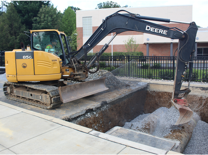Yellow Deere excavator digging a pit next to a sidewalk, against a building and fence.