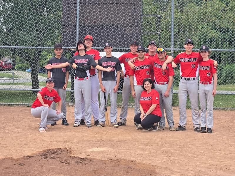 A group of baseball players and a coach in red and black uniforms pose for a photo on a dirt baseball field.