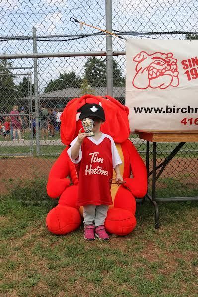 A small child in a Tim Hortons jersey and black hat stands holding a trophy in front of a large red plush dog mascot.
