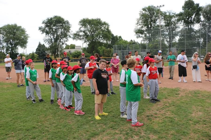 A group of children in baseball uniforms stand in a loose line on a grassy field with adults standing behind them.
