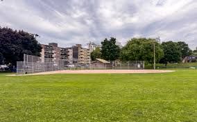 A baseball field with a chain-link backstop, sand infield, and green grass outfield under a cloudy sky.