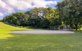 A baseball diamond with a sandy infield set in a green, grassy park surrounded by a backdrop of dense, leafy trees.
