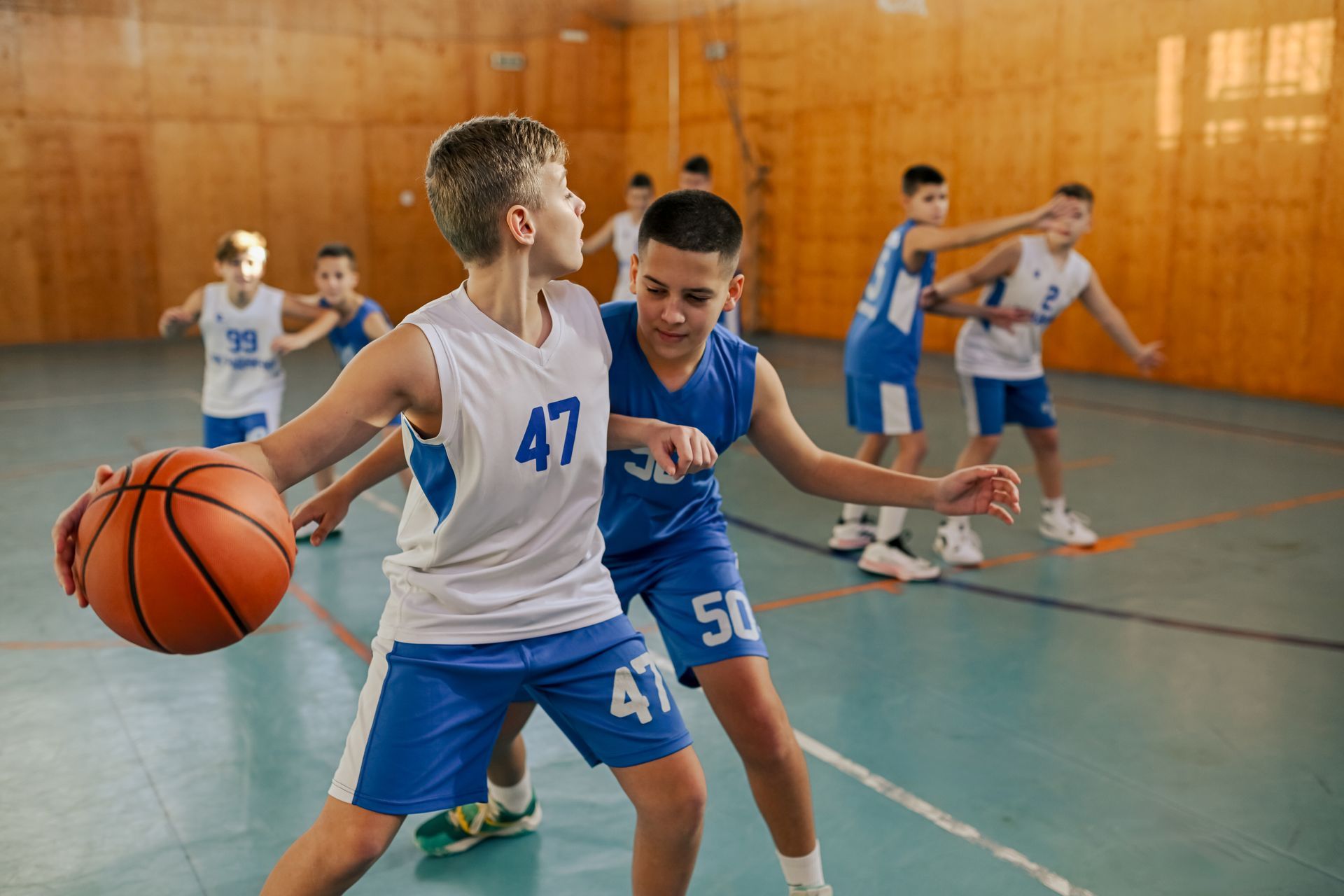 Youth basketball game in a gym, with a player dribbling while another defends nearby.