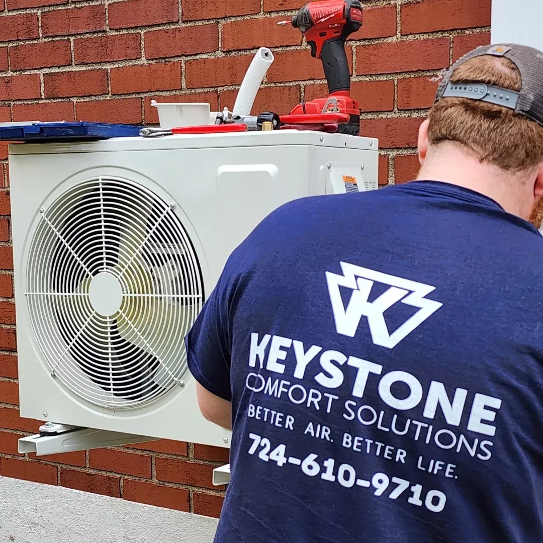 HVAC technician installing an air conditioning unit on a brick wall. The technician wears a blue shirt with the Keystone Comfort Solutions logo.