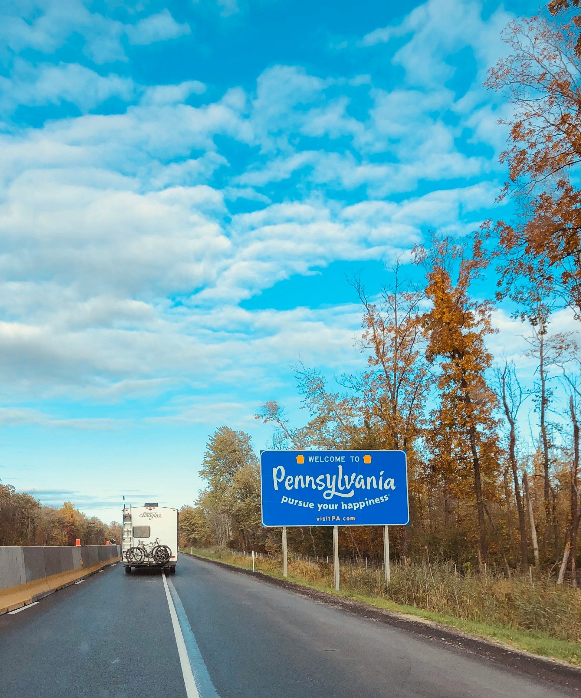 Pennsylvania state line sign on a sunny day. A vehicle approaches on a road lined with trees.