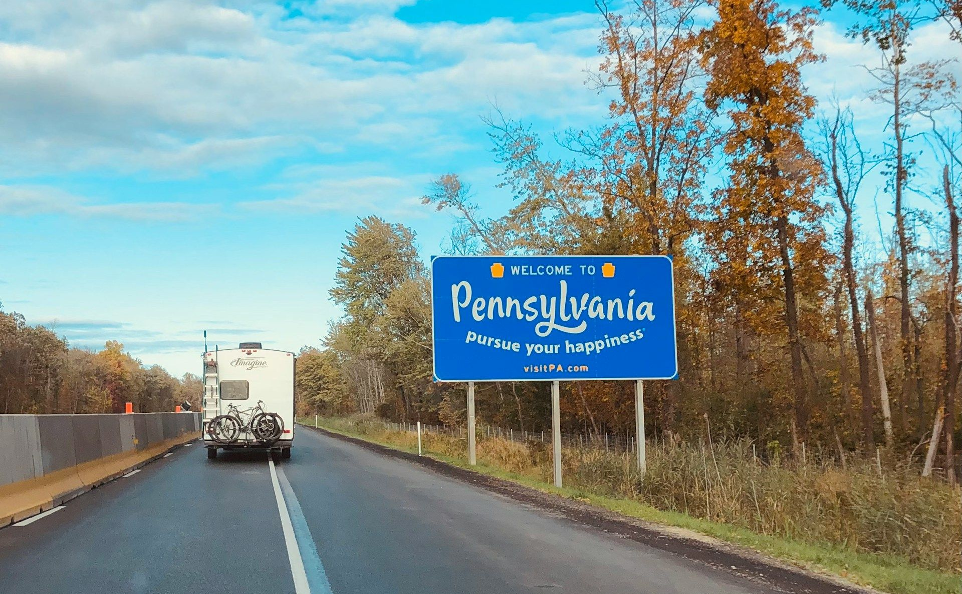 Pennsylvania state line sign on a sunny day. A vehicle approaches on a road lined with trees.