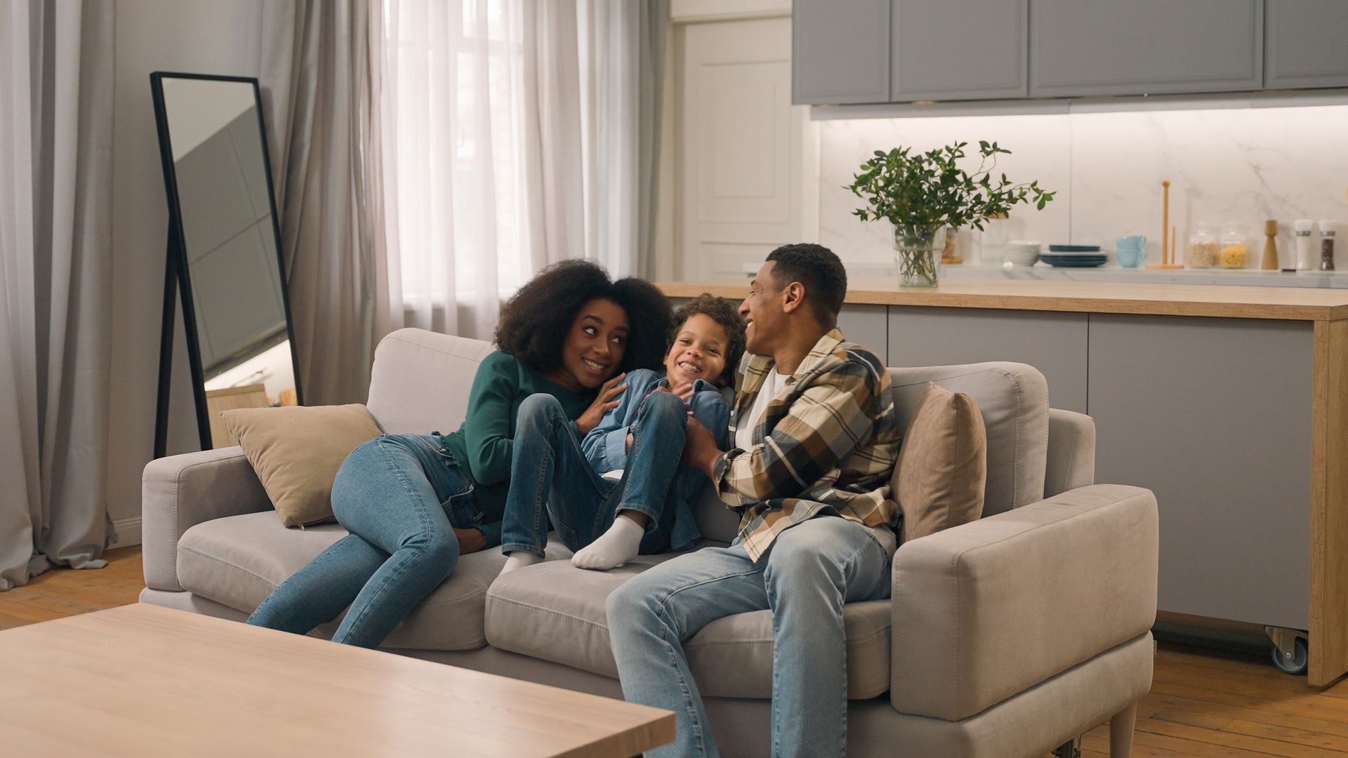 Family on couch, smiling. Living room with wood floor and kitchen in background.