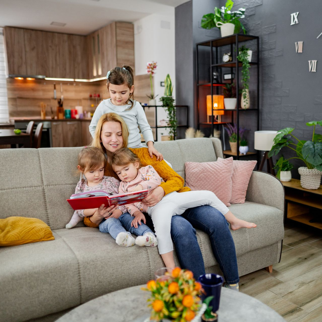 Woman reading with three children on a sofa in a living room; plants and decor visible.