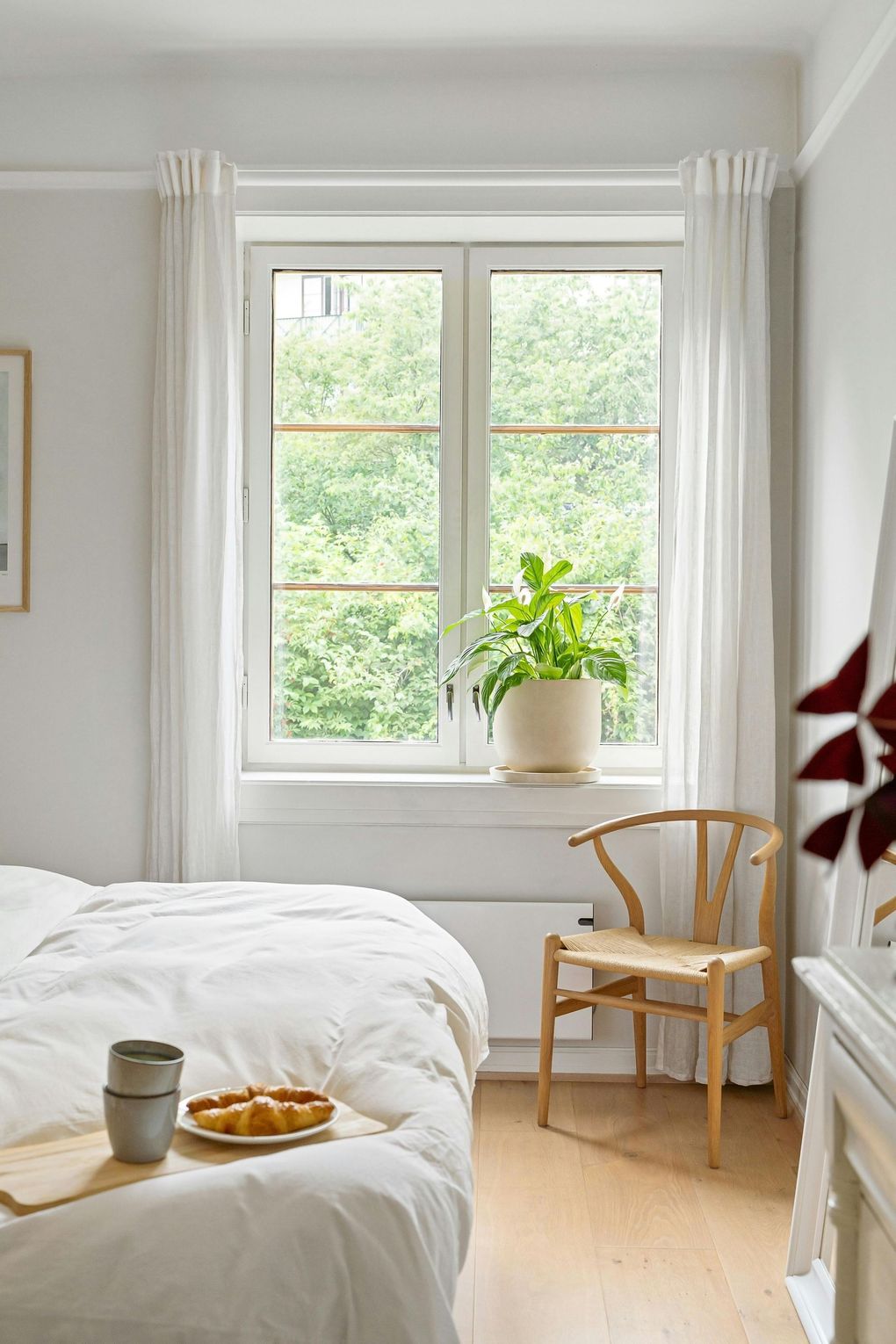 Lichte slaapkamer met een bed, een kleine stoel, een potplant bij het raam en een dienblad met ontbijt op bed.