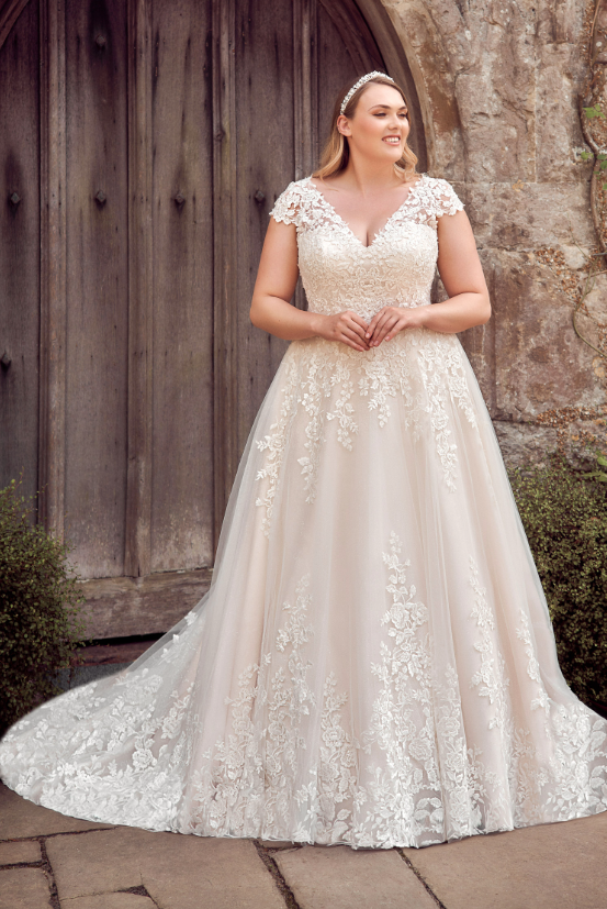 Curves wedding dress Envy by Phoenix PH8056 beaded glorious  floral lace create a v neckline and soft cap sleeve continuing from the fitted bodice onto the tulle skirt where a subtle glitter underlay sparkles gently. The v neckline continues to the back where a corset leads the eye to the chapel length train and full lace hem.