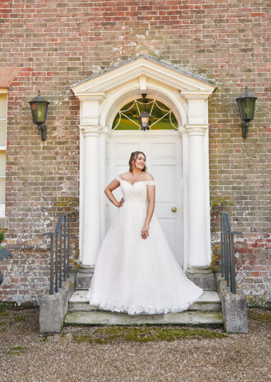 Envy by Phoenix PH7006 Softly curved off shoulder neckline with intricately beaded wild meadow flora lace applique that tumbles from the bodice to the full tulle skirt and onwards to the hem. The soft neckline continues to the back where a corset leads the eye to the chapel train.