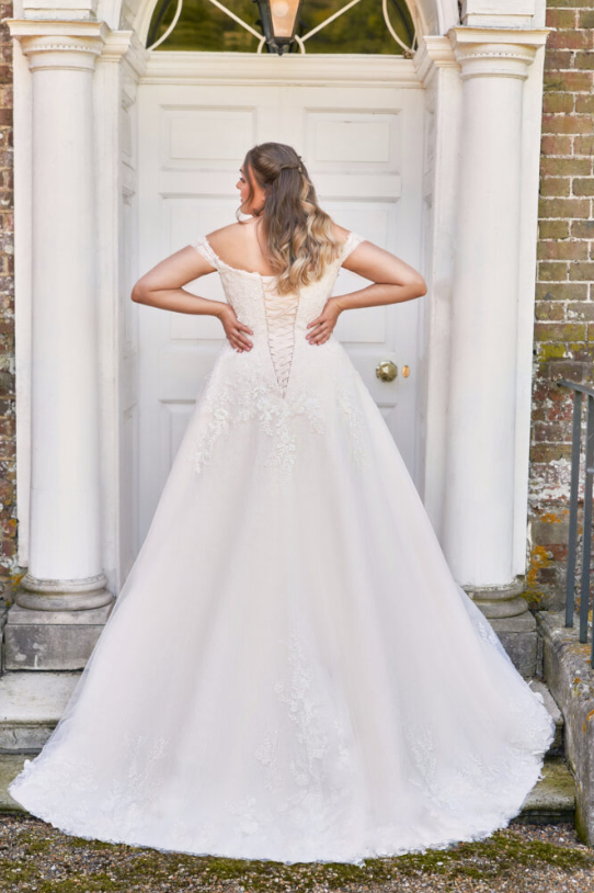 Envy by Phoenix PH7006 Softly curved off shoulder neckline with intricately beaded wild meadow flora lace applique that tumbles from the bodice to the full tulle skirt and onwards to the hem. The soft neckline continues to the back where a corset leads the eye to the chapel train.