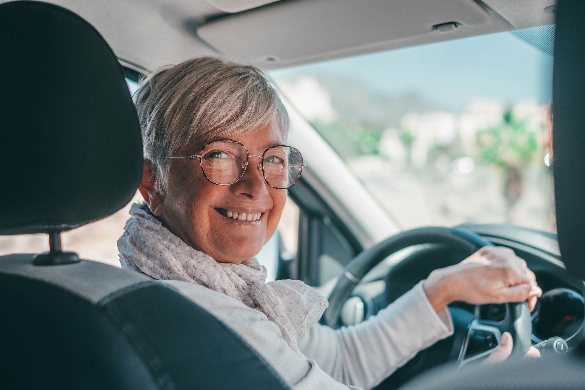 A person smiling while driving a car, wearing round glasses and a light-colored scarf, seen from the backseat.