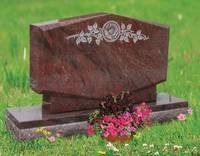Brown granite gravestone with pink flowers at its base on a grassy cemetery lawn