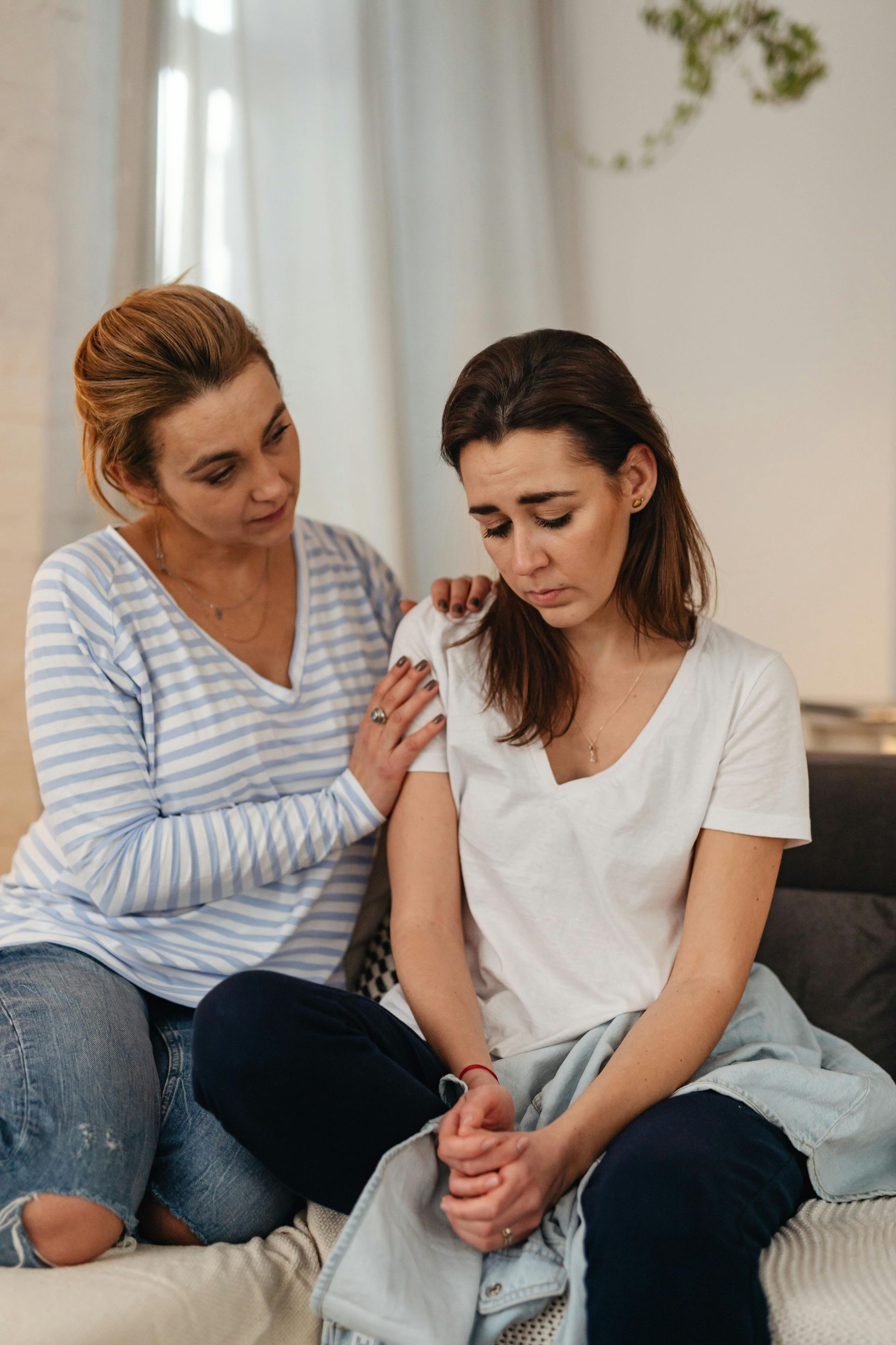 Two women sit on a couch; one comforts the other, who looks upset.