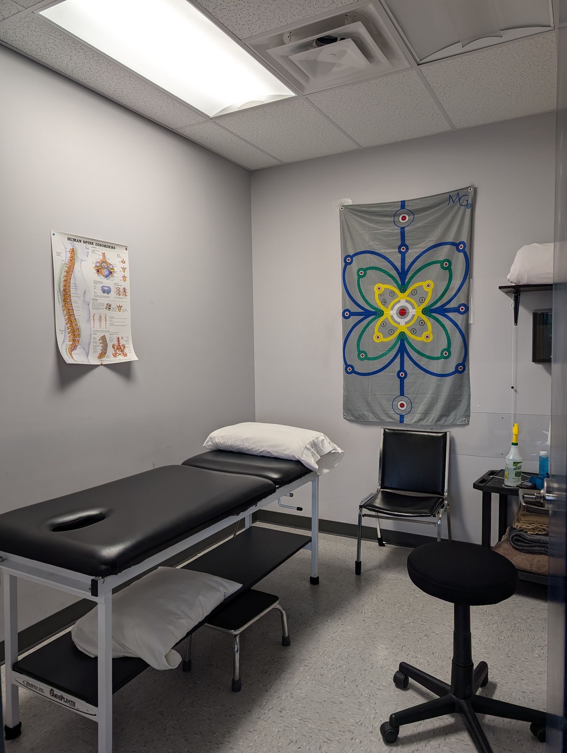 Exam room with exam table, chair, stool, and colorful wall art under fluorescent lights
