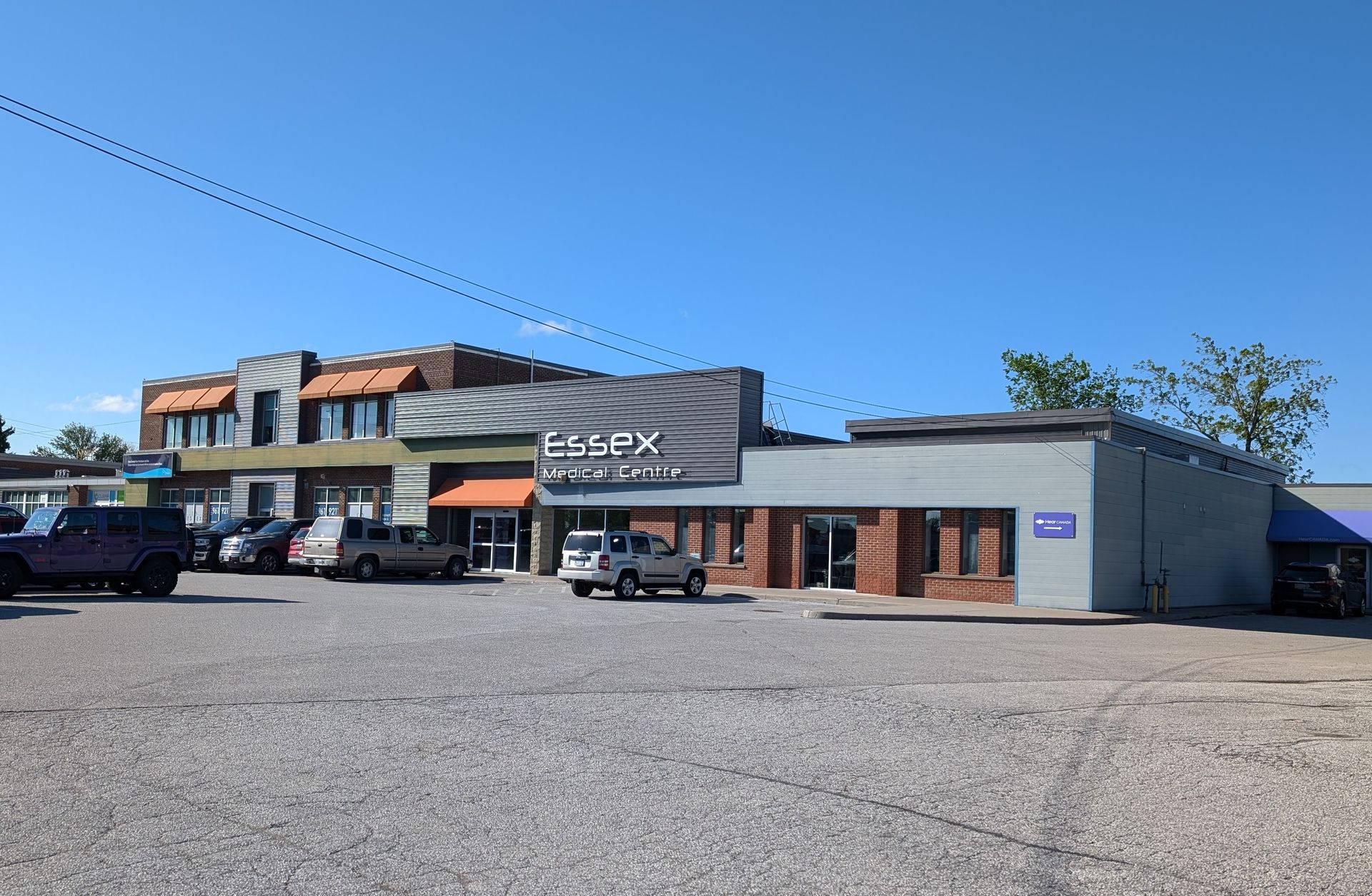 Small strip mall with parked cars under a clear blue sky, featuring a central brick storefront and gray building wing
