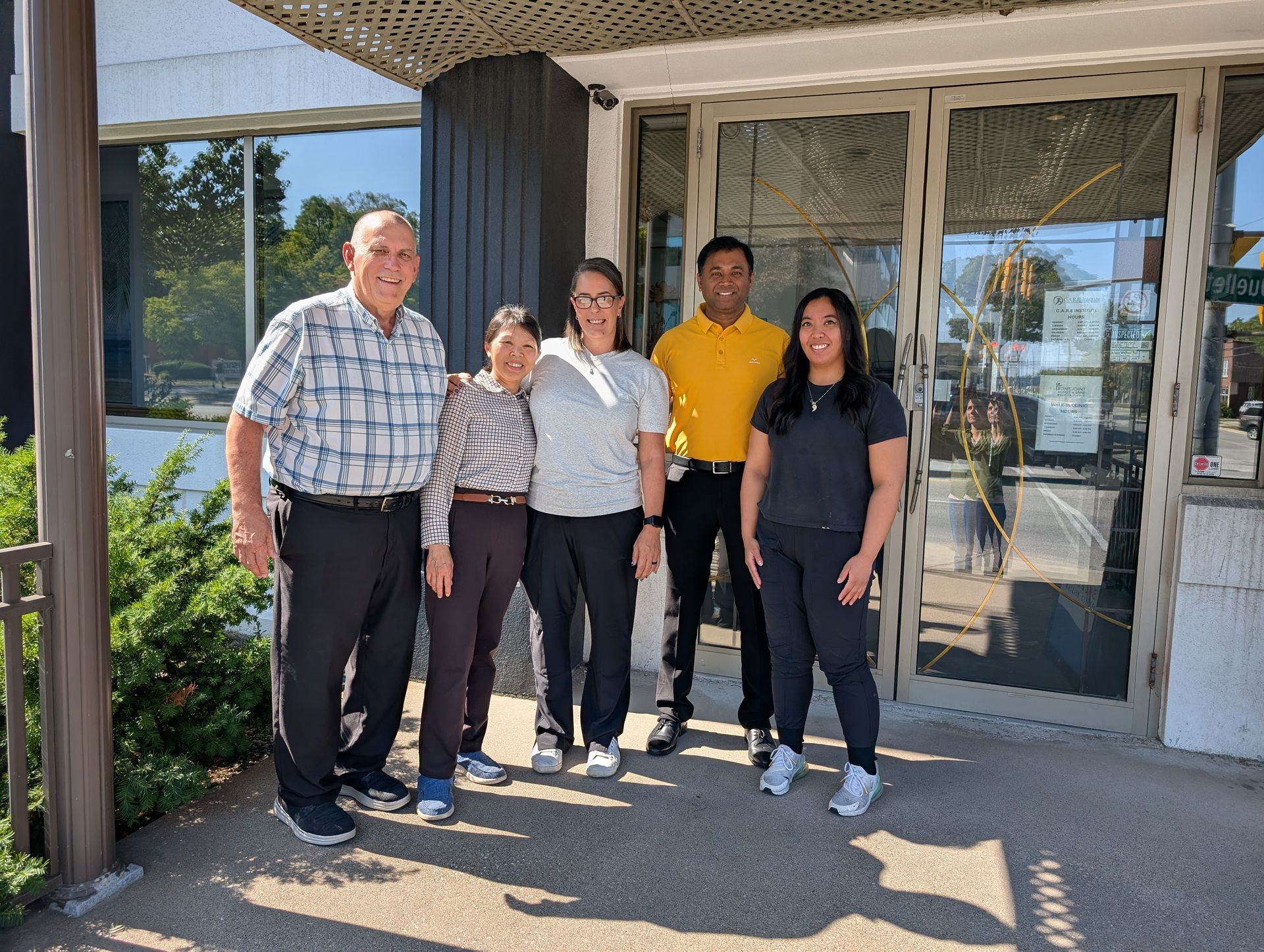 Five people stand smiling outside a building entrance in bright sunlight.