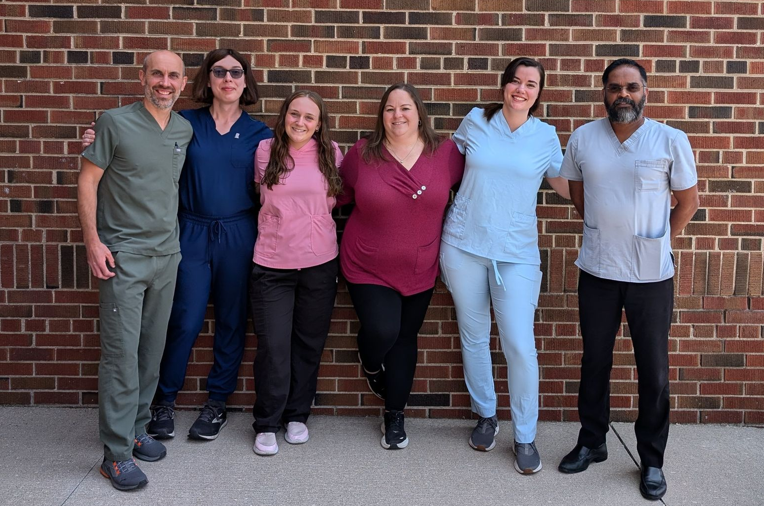 Six people standing shoulder to shoulder outdoors against a brick wall, smiling at the camera.