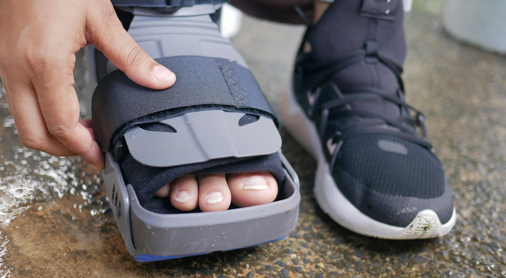 A hand adjusts a black toe-cover sandal on a foot beside a black sneaker on wet pavement.
