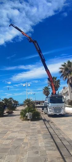 Un camion che solleva un lungo braccio rosso stagliandosi contro un cielo azzurro brillante su una strada lastricata in pietra.