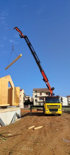 Cantiere edile con una gru che solleva una lunga trave accanto a un camion giallo sotto un cielo azzurro e limpido.