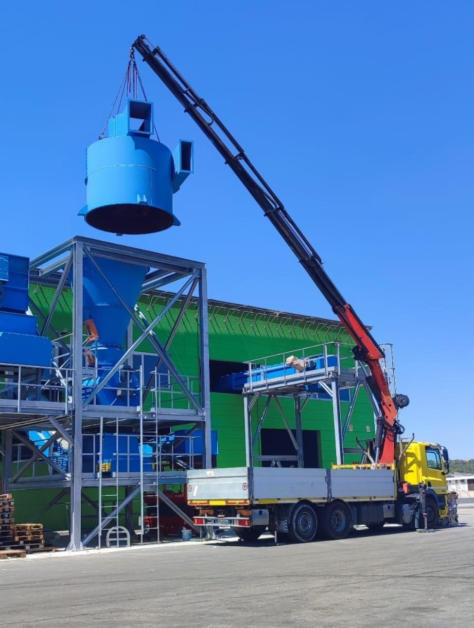Una gru solleva un serbatoio industriale blu accanto a un camion in un cantiere.