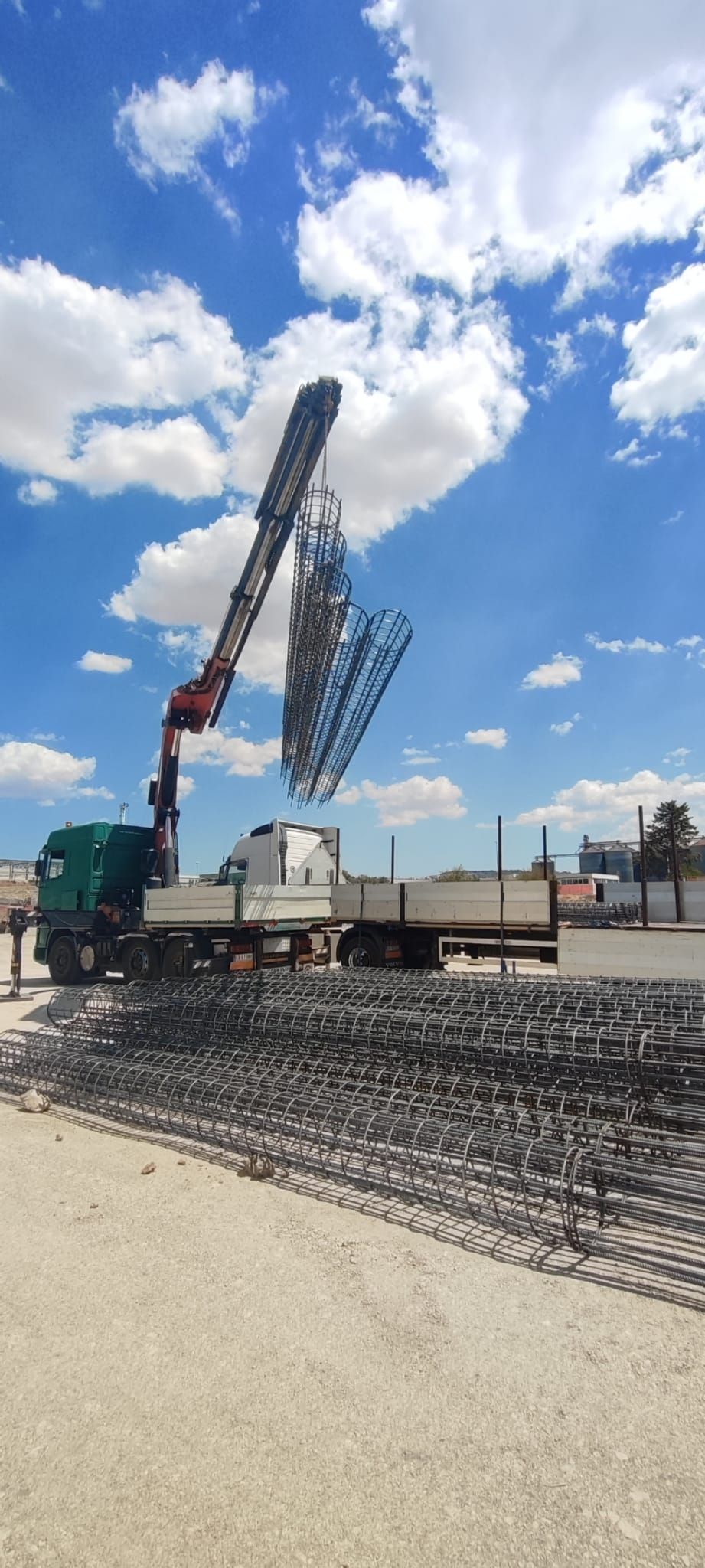Una gru da costruzione solleva una trave d'acciaio in un cantiere sotto un cielo azzurro brillante con nuvole.