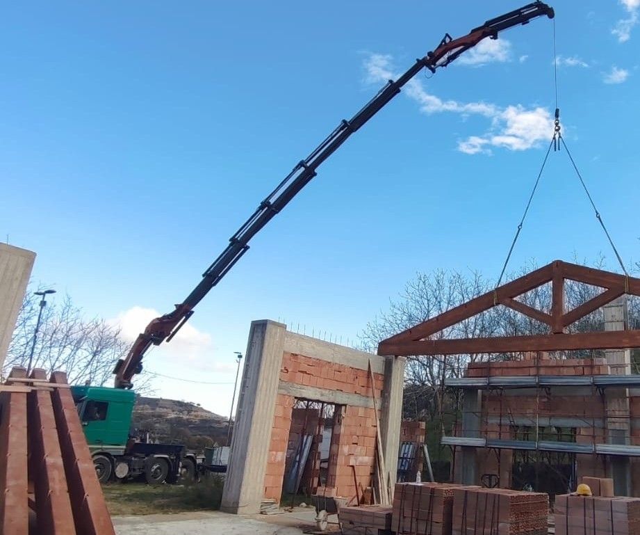 Cantiere edile con una gru che solleva una struttura di tetto in legno sullo sfondo di un cielo azzurro.