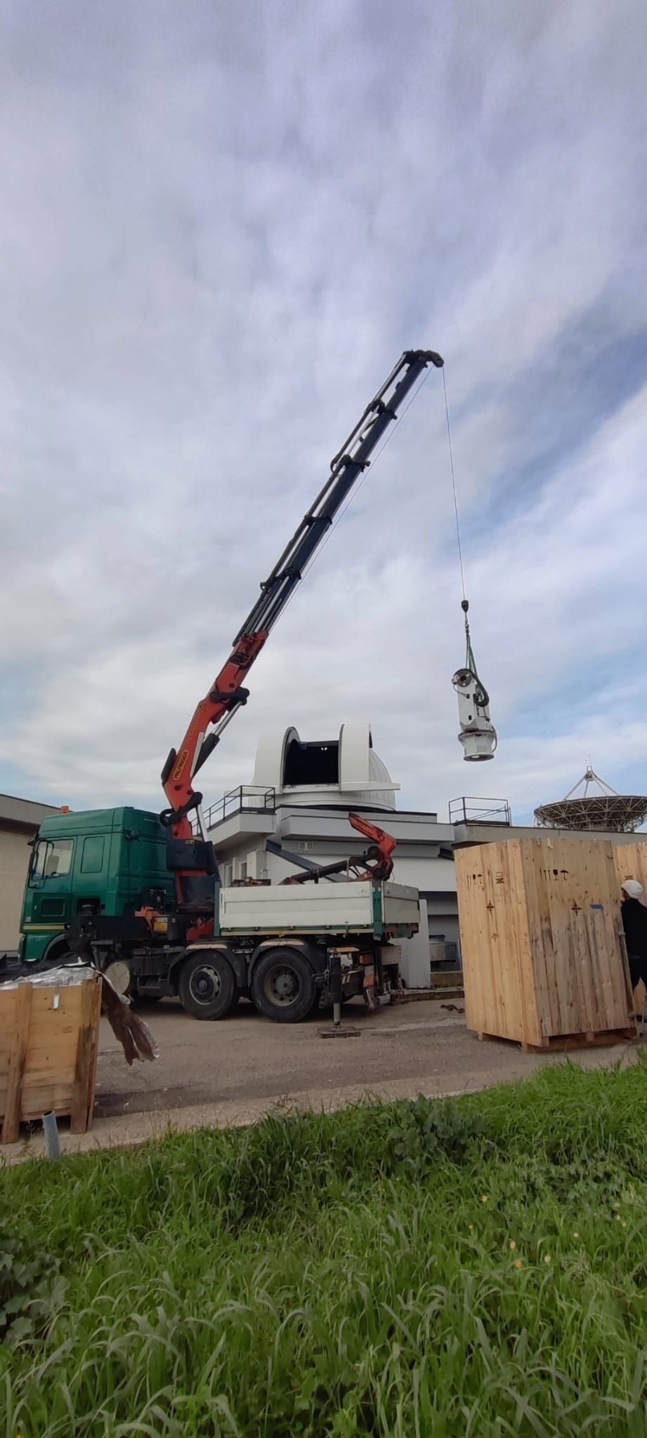 Un camion gru solleva un lungo braccio accanto a una cassa di legno in un cantiere edile sotto un cielo nuvoloso.