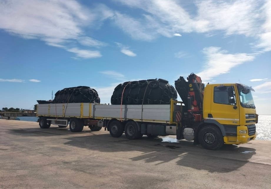 Un camion giallo con due rimorchi carichi di merci coperte è parcheggiato su un'ampia area pavimentata sotto un cielo azzurro.