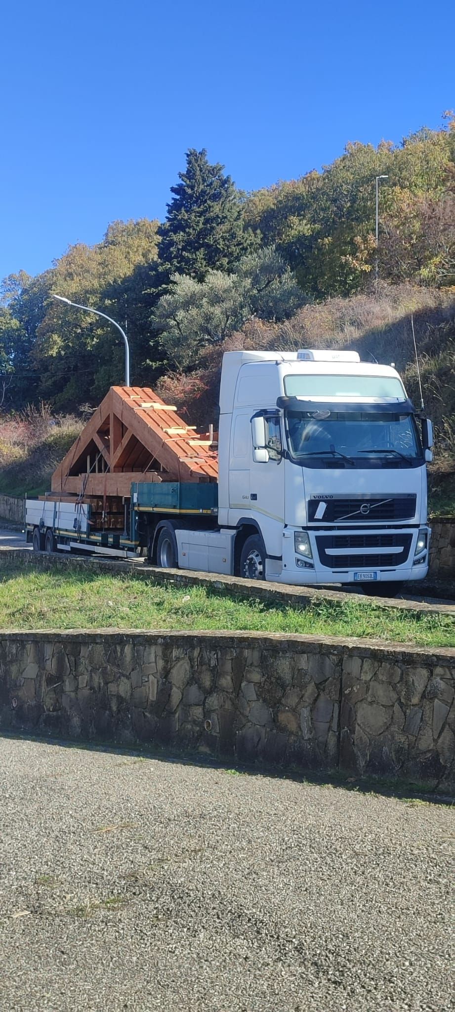 Un camion bianco che trasporta macchinari arancioni è fermo a bordo strada, accanto ad alberi e un muro di pietra.