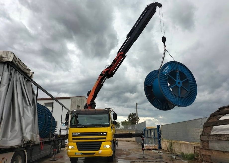 Un camion gru giallo solleva una grande bobina di cavo blu in un piazzale industriale sotto un cielo nuvoloso.
