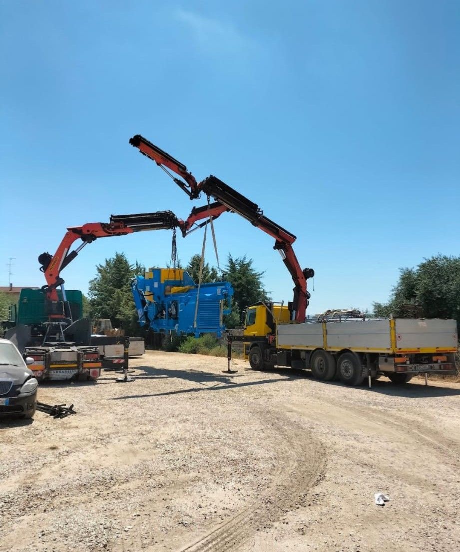 Cantiere edile con una macchina blu e una gru rossa che sollevano dei camion su un piazzale ghiaioso.