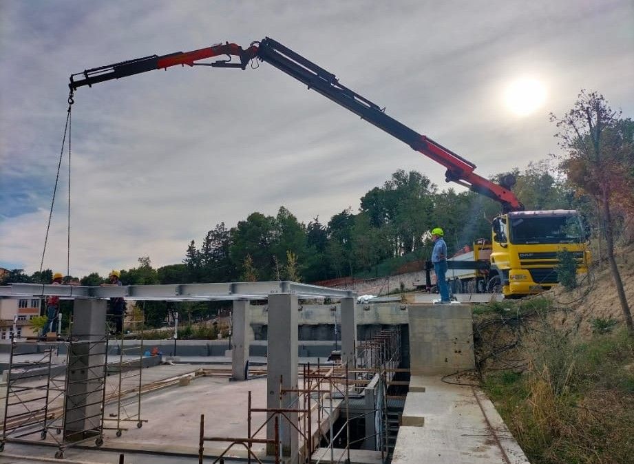 Una gru sta versando cemento in un cantiere edile accanto a un ponte, con operai e macchinari ben visibili.
