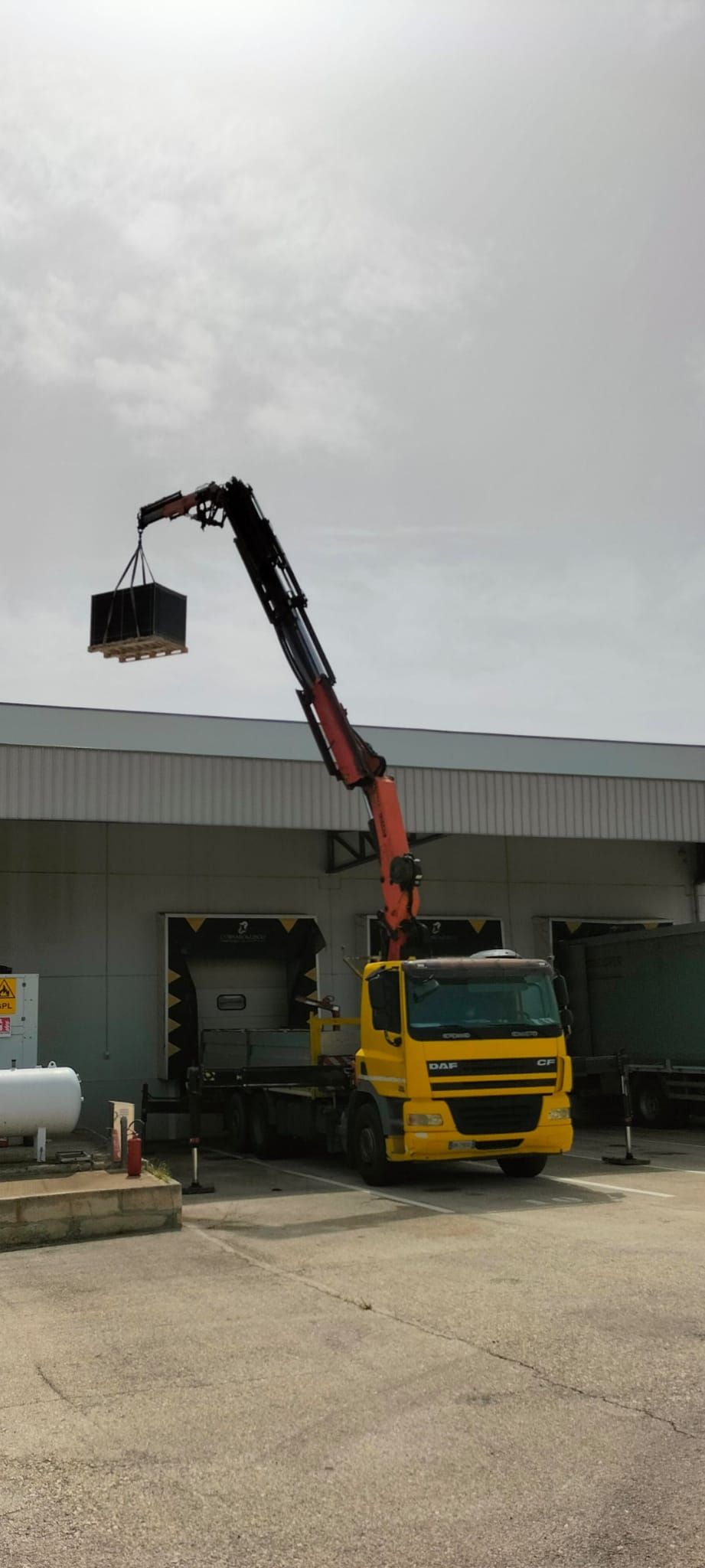 Un camioncino giallo con gru sollevata che carica un container nero all'esterno di un magazzino.