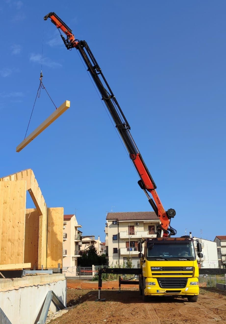 Un camion con gru solleva una lunga trave di legno in un cantiere edile sotto un cielo azzurro e limpido.