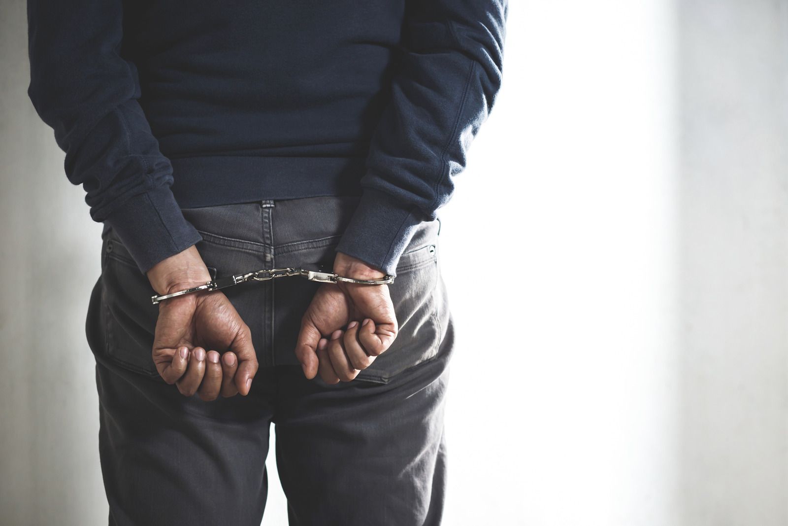 Person in handcuffs, viewed from the back, against a light wall.