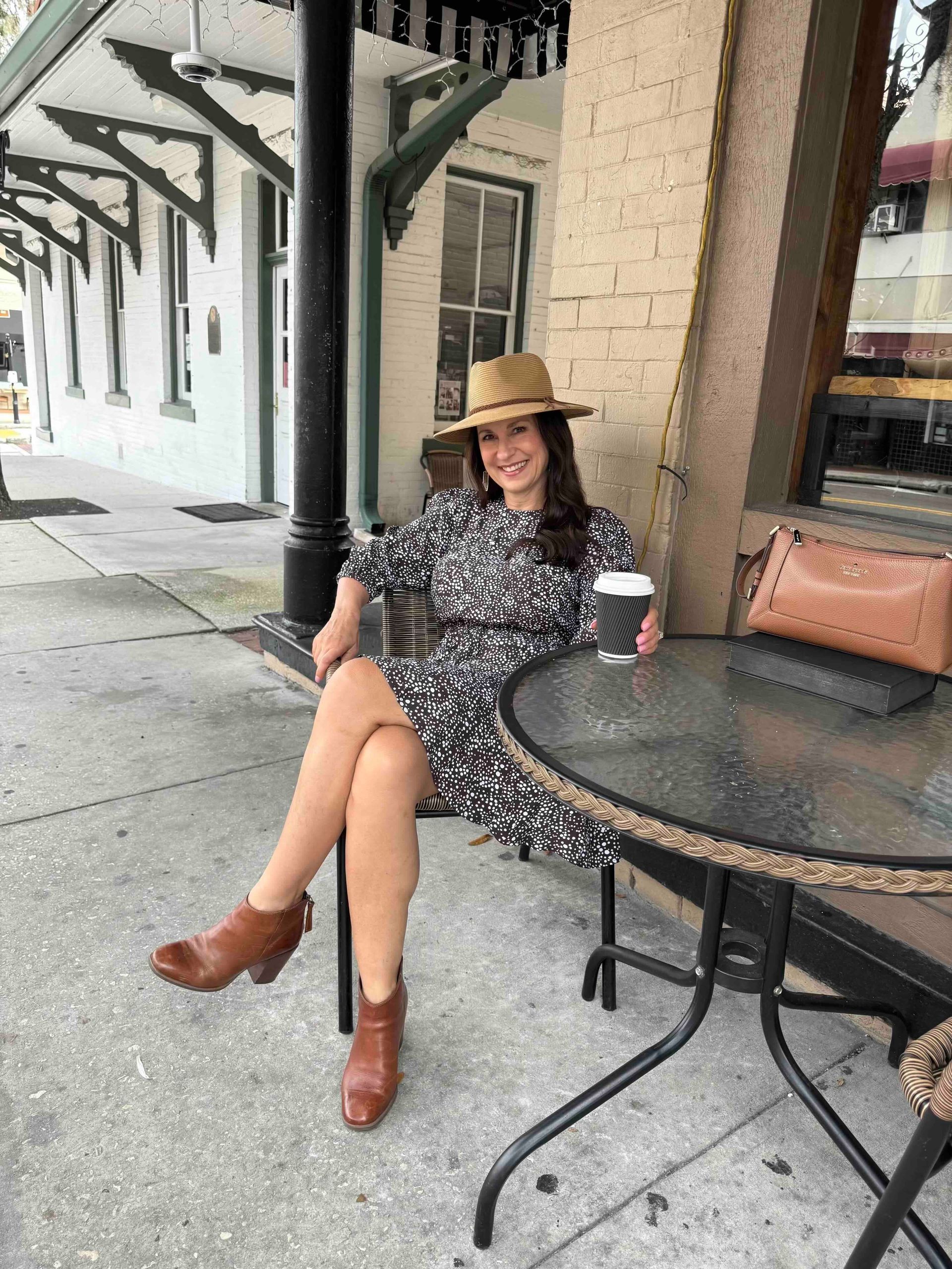 Woman in floral dress and hat sits at outdoor cafe table, holding coffee, brown boots.