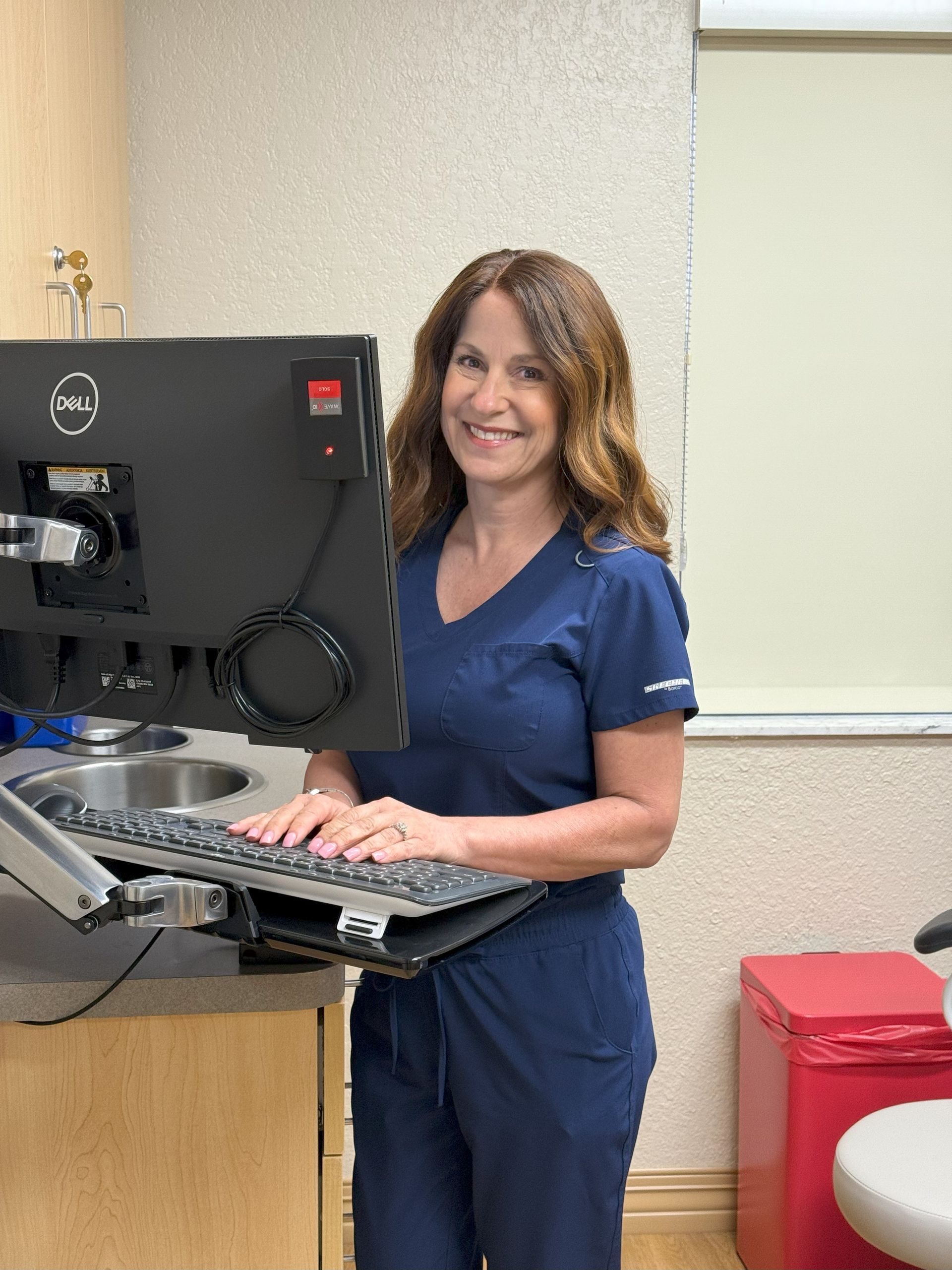Woman in blue scrubs at a computer, smiling. Medical office setting with sink and red disposal bin.