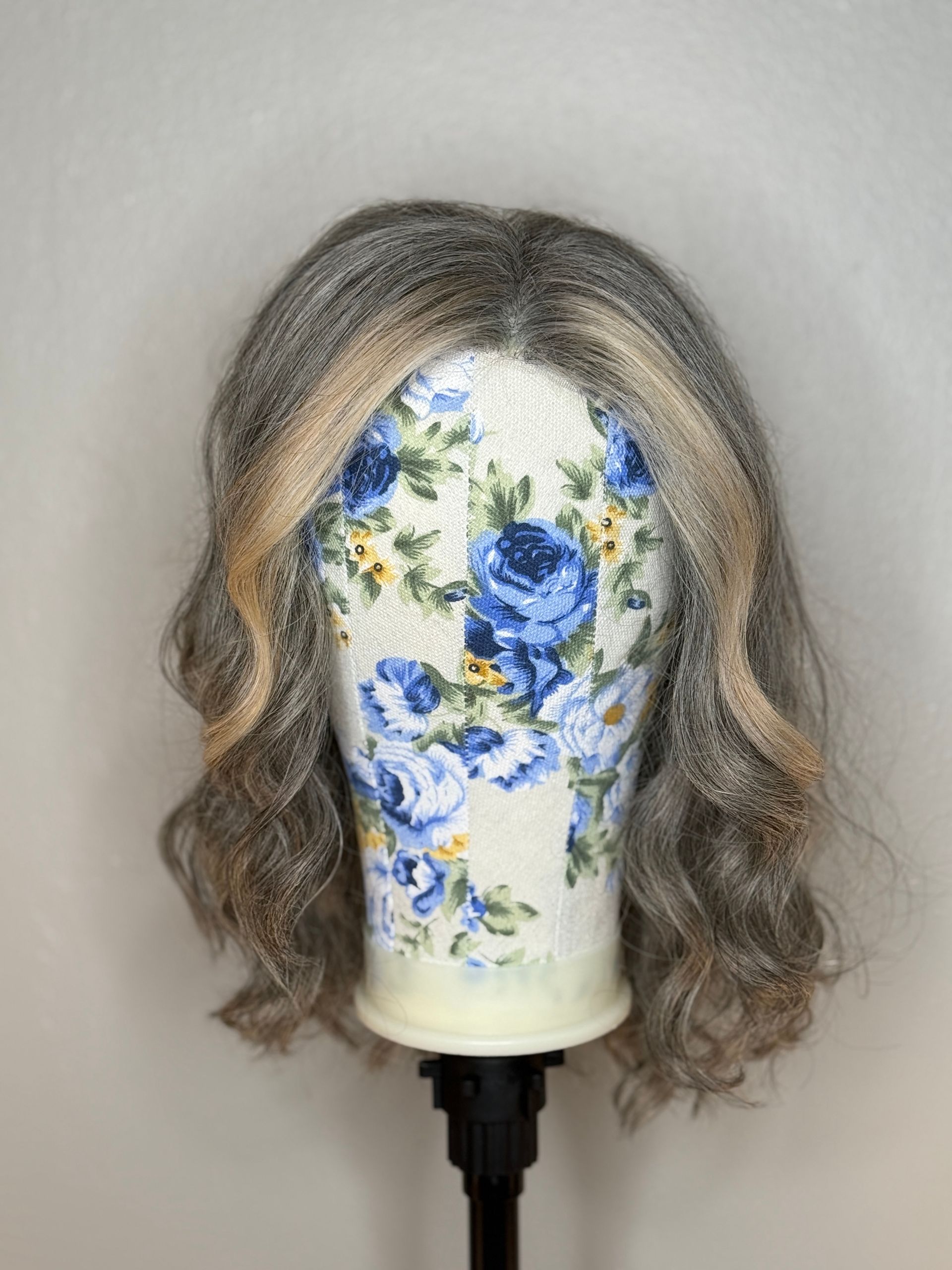 Gray wig on a floral-covered wig stand. The wig has blonde highlights and loose curls.