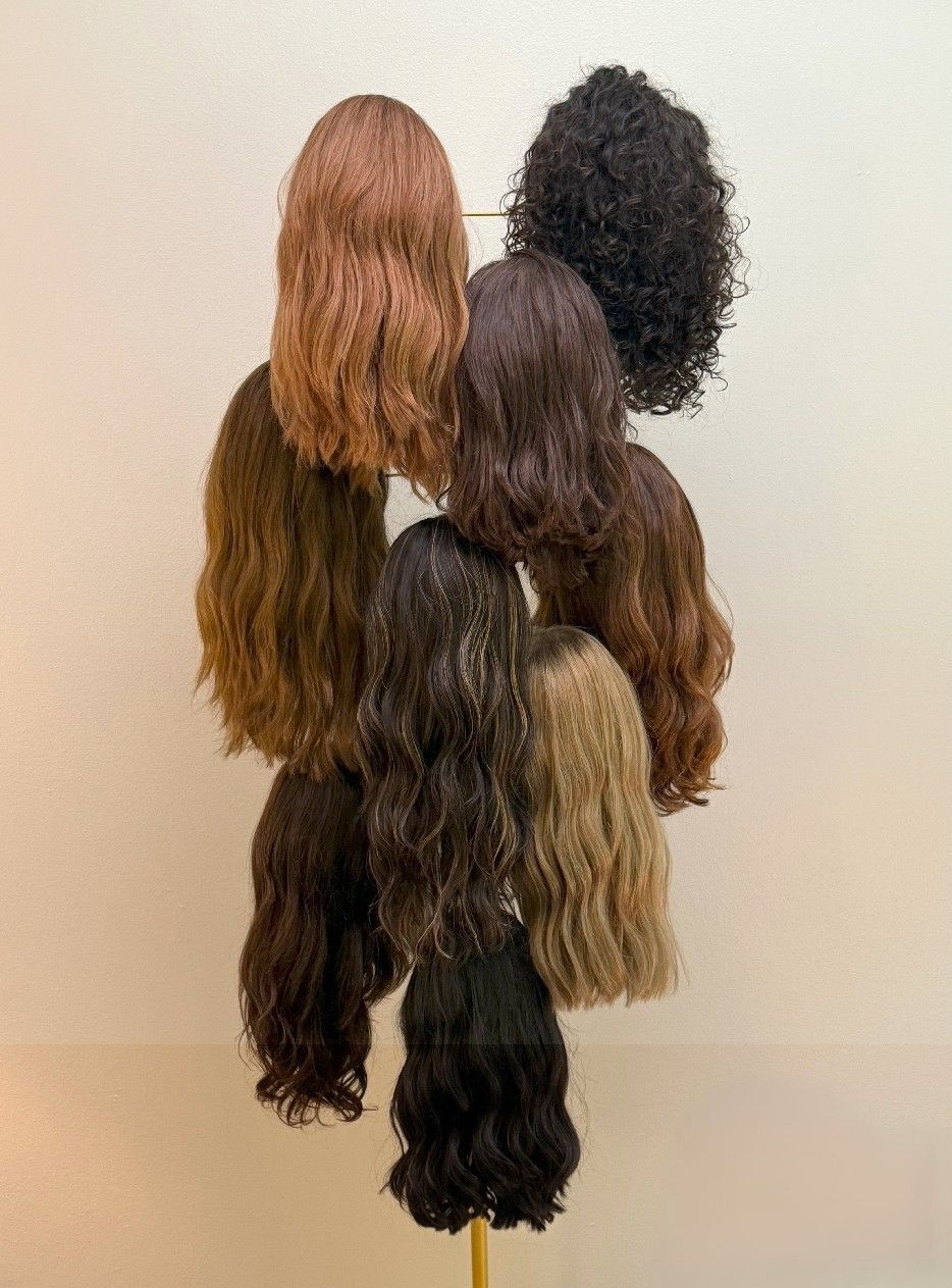 Wigs of various shades of brown and black hanging on a gold stand against a cream wall.