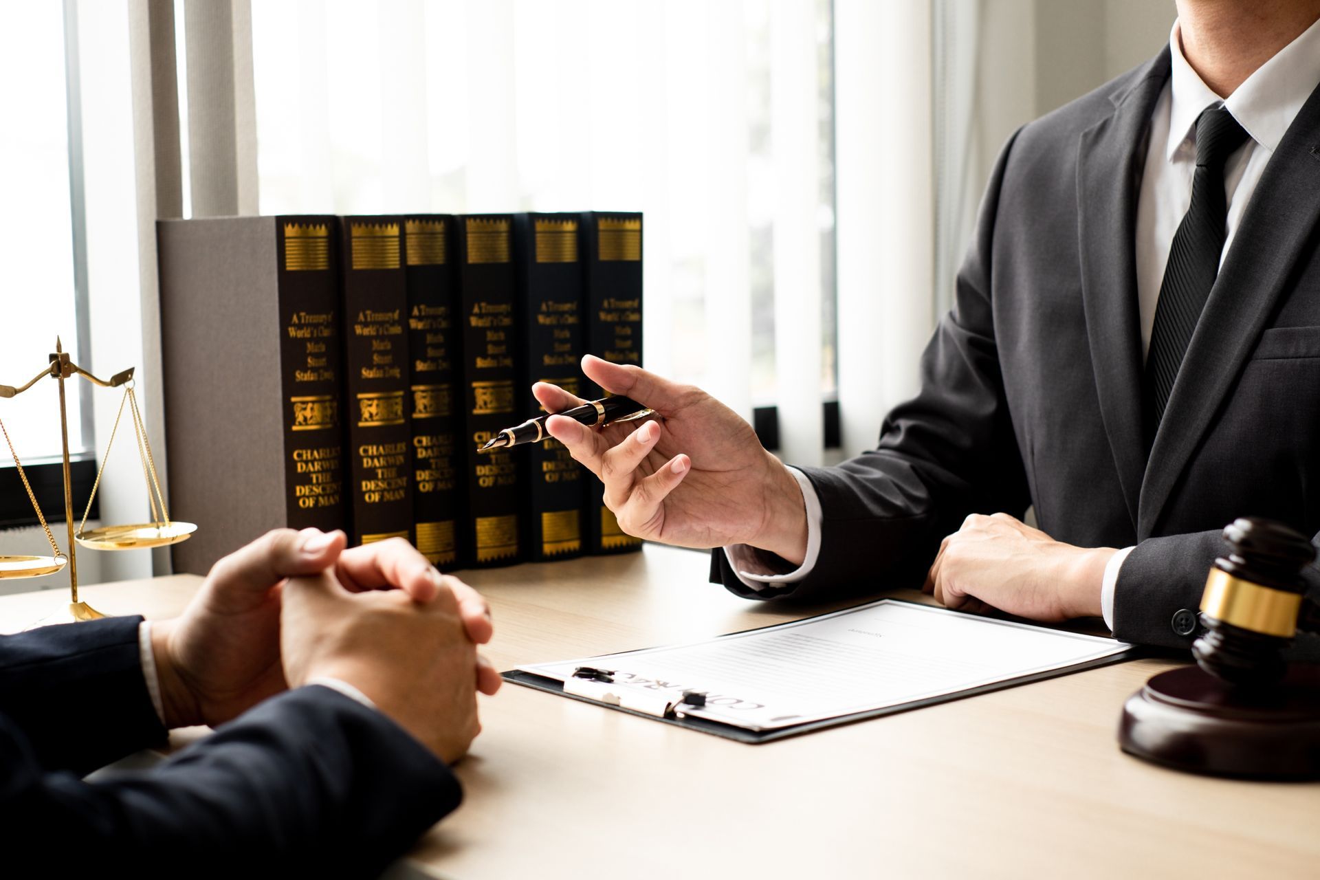 Lawyer in suit gestures while speaking to client at desk with documents and law books. Lawyer in suit gestures while speaking to client at desk with documents and law books.