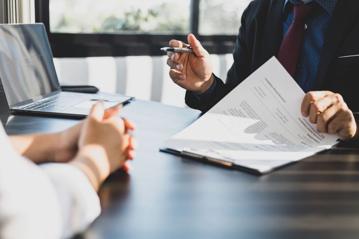 A professional sitting at a desk holds documents while gesturing during a meeting with an unseen person.