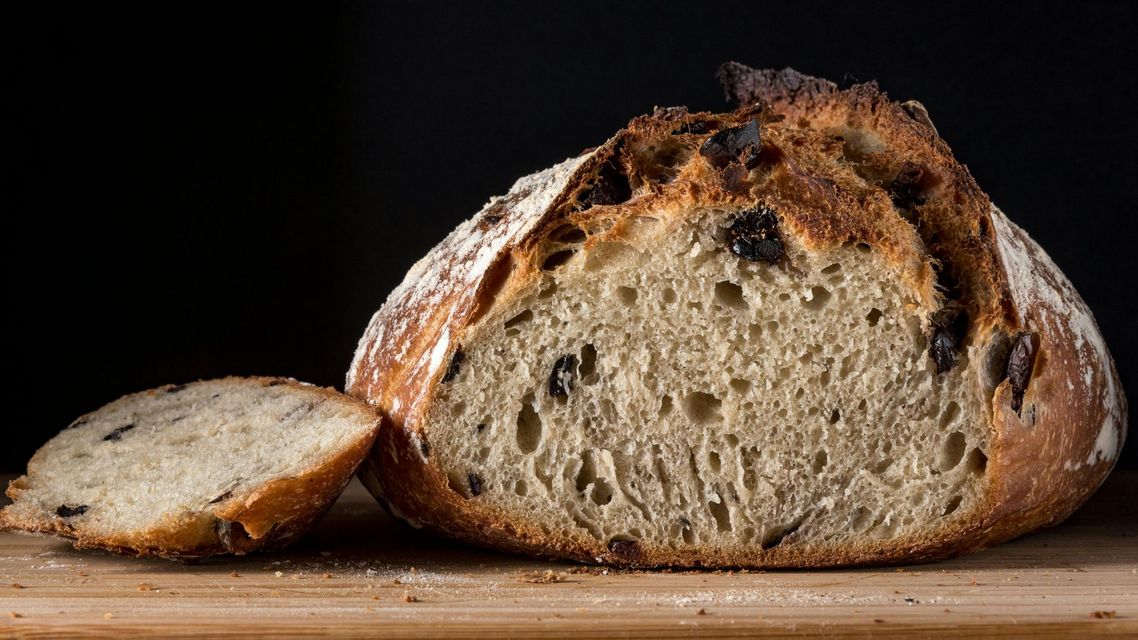 Pagnotta di pane tagliata a metà, con inclusioni di olive; su una superficie di legno, sfondo nero.