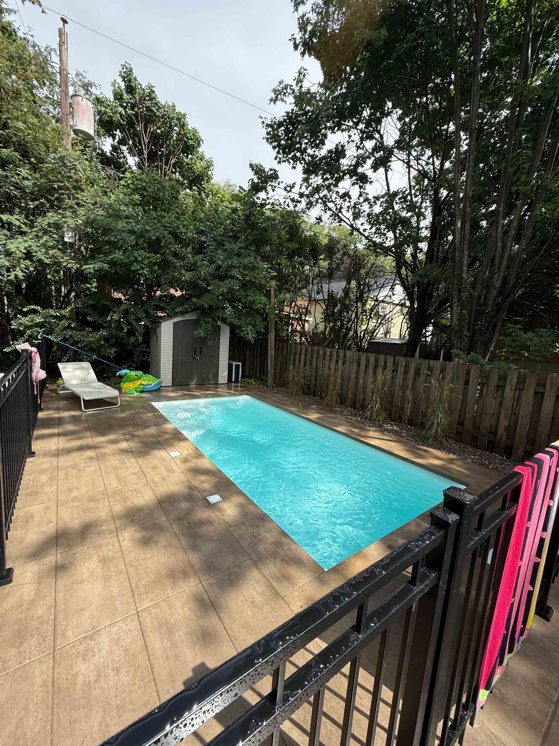 Piscine dans le jardin entourée d'arbres et d'une terrasse, avec une clôture noire au premier plan.